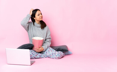 Young mixed race woman eating popcorn while watching a movie on the laptop having doubts while scratching head