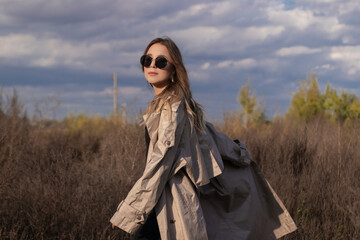 brunette in fashionable trench coat, black dress and sunglasses in the field. autumn, yellow leaves, blue sky.