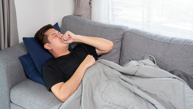 Asian man with seasonal infections Cold Blowing His Nose and sneezing into Tissue with headache lying on sofa with high fever and a flu