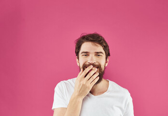 Happy young man on a pink background in a white T-shirt and a thick beard gesticulate with his hands