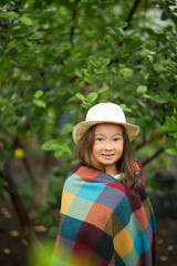 portrait of adorable asian little girl around plants, child is wrapped in plaid, wearing hat, look at camera. in the garden