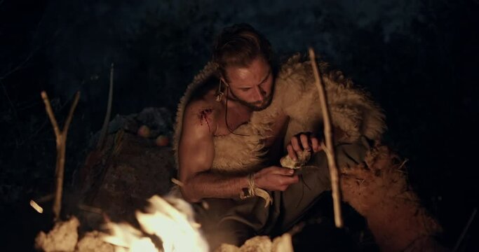 Primeval man near bonfire in forest. Prehistoric male hunter wearing fur clothes sitting at bonfire and sharpening handmade knife with stone at night in woods
