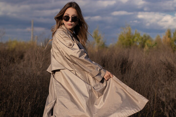 brunette in fashionable trench coat, black dress and sunglasses in the field. autumn, yellow leaves, blue sky.