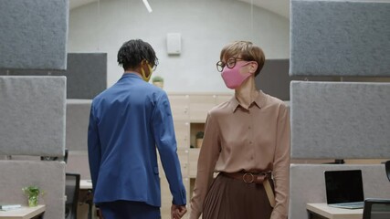 Medium shot of young African-American businessman and his female Caucasian coworker both wearing masks greeting each other in office. Woman sitting down at workplace and starting working