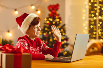 Happy child waving hand at laptop screen greeting his friends at online Christmas party