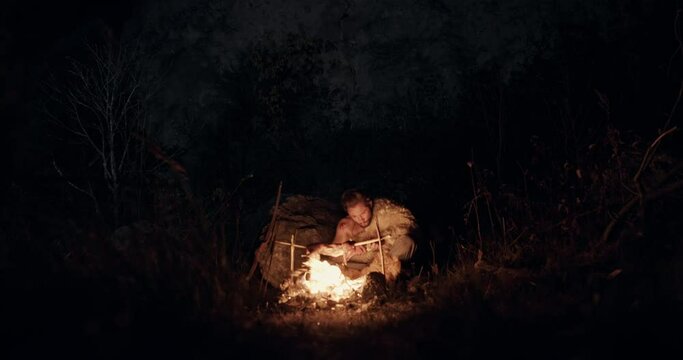 Savage man roasting meat over bonfire. Lonely savage male hunter sitting in front of burning bonfire with killed animal roasted over flame in dark forest at night