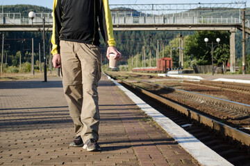 Young man in sport tourist clothes stay on the platform of the station, near the rails, and holds a cup of coffee