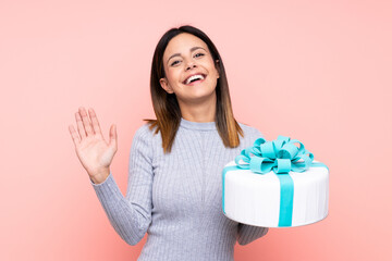 Woman holding a big cake over isolated pink background saluting with hand with happy expression