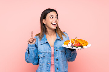 Brunette woman holding waffles over isolated pink background intending to realizes the solution while lifting a finger up