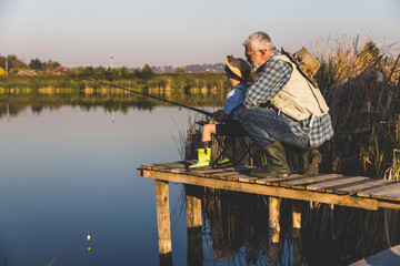 Senior man sitting on jetty fishing in lake with grandson