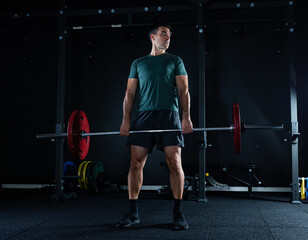 Fototapeta na wymiar Tall dark man with green t shirt lifting barbell with red weights on a dark gym.