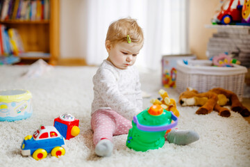 Happy joyful baby girl playing with different colorful toys at home. Adorable healthy toddler child having fun with playing alone. Active leisure indoors, nursery or playschool.