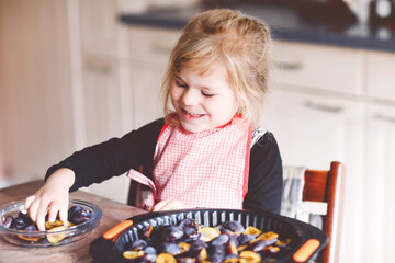 Cute little toddler girl baking plum pie at home. Happy smiling child helping and preparing plums...