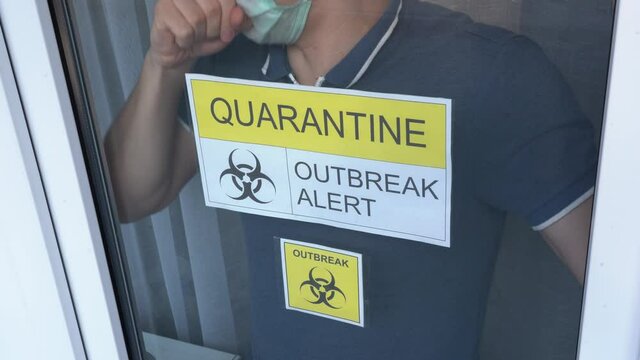 Sick isolated man in blue T-shirt leans against window glass with yellow warning inscription QUARANTINE OUTBREAK ALERT. Guy wear face mask, coughs, looks out of hospital medical room close up. No face