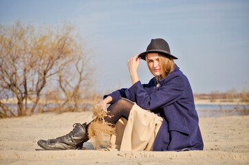 Young caucasian blonde girl in a light dress, coat and hat on beach, under rays of spring sun with reeds branches