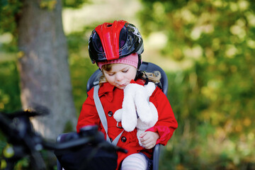 Little toddler girl with security helmet on head sitting in bike seat of her mother or father bicycle. Safe and child protection concept. Family ctivity trip on autumn day. Kid holding push soft toy