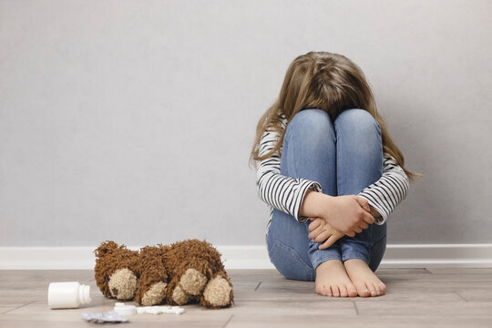 Sad teenager girl, schoolgirl in depression sits on the floor near the wall, next to a teddy bear and pills.