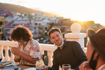 Young cheerful people enjoy dinner outdoor on patio while wearing surgical face mask under chin - Sunset in the background