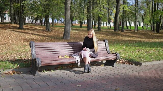 A young blonde girl with long hair sits on a bench and walks in a city park on a sunny day in autumn