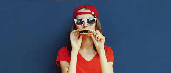 Portrait close up of young woman eating burger wearing baseball cap, sunglasses over blue background