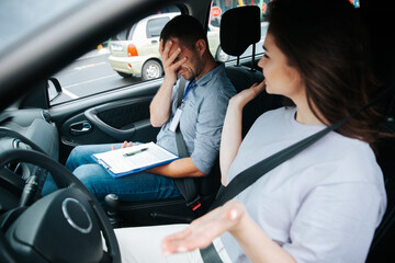 Attractive female driver in white shirt turns her head to upset male auto instructor making facepalm gesture. Instructor on driving courses is stressed with student's mistakes.