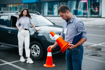 Male tired driving instructor collects traffic cones at the end of the work shift. Blurred female in light casual clothes student at background standing near the black car. Wet asphalt after rain.