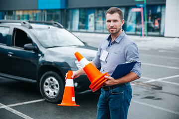 Driving instructor loves his job. Young attractive man standing with clipboard and cones in his hands and looking at camera. Black car at background. Obtaining a driver's license concept.