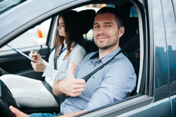 Happy male student showing thumbs up and smiling. Safe driving concept. Cheerful young man excited about his successful driving license exam. Female instructor wrtiting results of test in her paper.