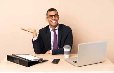 Young business man in his office with a laptop and other documents holding copyspace imaginary on the palm to insert an ad