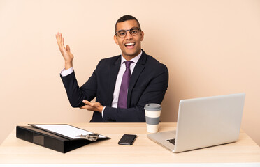 Young business man in his office with a laptop and other documents extending hands to the side for inviting to come