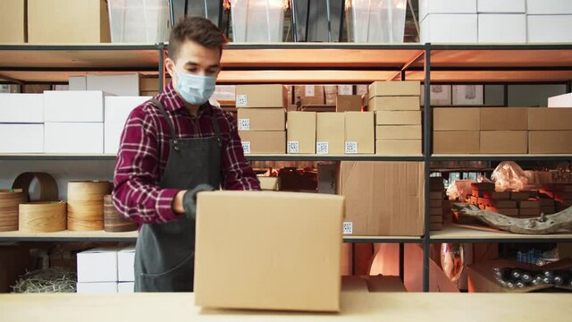 Male workers wearing protective face mask working in factory warehouse. Latin amrican man carrying box parcel walking indoor during covid 19 pandemic crisis. Logistic online shopping industry concept.