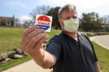 Man wearing a face mask holding out an I voted today sticker at a polling location