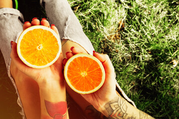A tattooed hipster girl in torn jeans with red nails sits on the grass and holds halves of a juicy orange Hands close up Diet concept