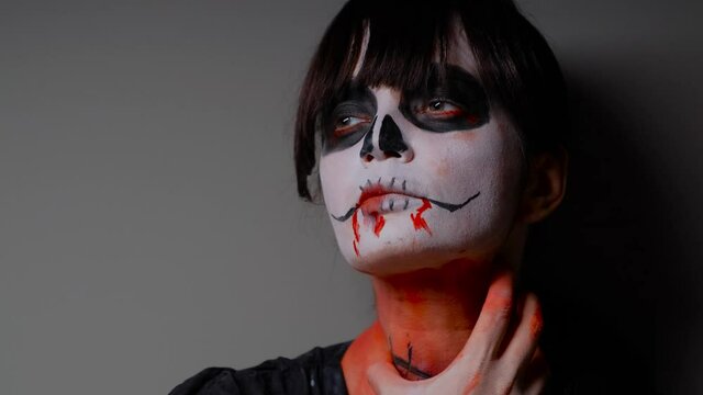 A close-up of the face of a zombie girl with dark bangs, who stands against the wall and holds her scarred throat with her painted hand. Halloween Eve.