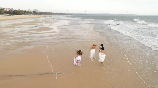 Beautiful girls in wedding dresses run along the beach on the water, the drone flies after them. Slow motion aerial video of happy brides on the sea coast