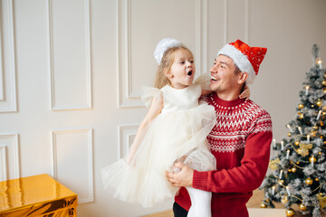 Fototapeta premium A man in New Year's clothes holds his daughter in his arms.Christmas concept, family holiday. Gift boxes and christmas tree