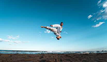 Black belt karate fighter giving a flying kick on the beach at sunset.