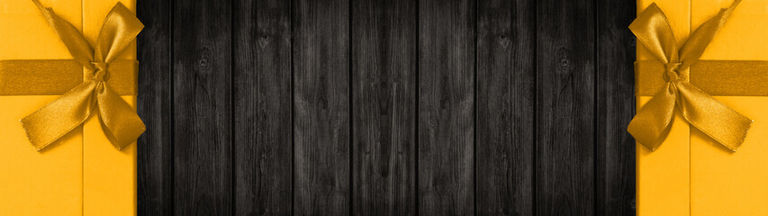 Naklejka premium Festive christmas / celebration / birthday background banner panorama - Yellow gift boxes with golden ribbon, isolated on dark black rustic wooden boards wall / wood texture, with space for text