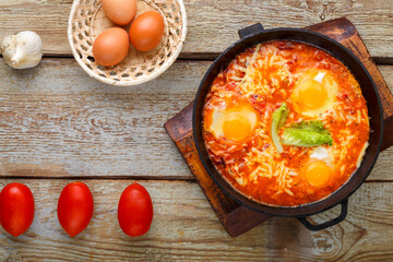 israeli fried eggs shakshuka with cheese in a frying pan near eggs and tomatoes.