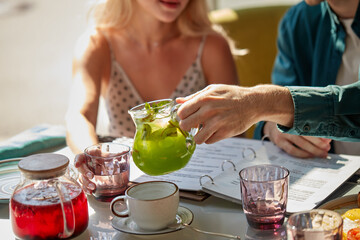 young gallant man pours sweet juice beverage into glasses for women, treats them. in cafe,...
