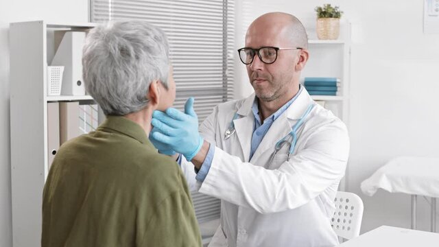 Over-the-shoulder of Caucasian male therapist sitting in medical office in front of unrecognizable grey-haired patient, palpating her neck and back of head with hands in gloves