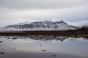 Snow covered mountain reflecting in water of black sand beach in Iceland