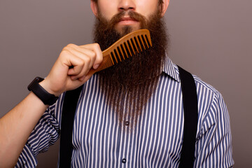 Cropped shot of stylish serious man with long beard. Man wearing shirt holding comb while standing...