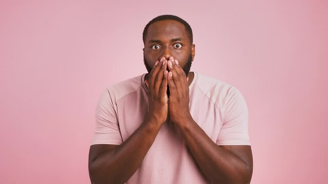 Shocked black man feeling excitement, looking at camera and holding hands near face, pink background