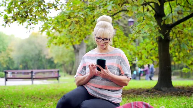Portrait happy senior woman sitting in park on the lawn using smartphone or phone in hands. Mature female uses app in gadget in nature outdors. emotionally browsing news and photos on social network