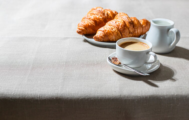 Croissants and cup of coffee in the morning light, sunlight shadow, on a linen fabric. Morning breakfast concept.