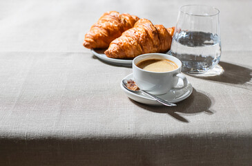 Croissants and cup of coffee in the morning light, sunlight shadow, on a linen fabric. Morning breakfast concept.