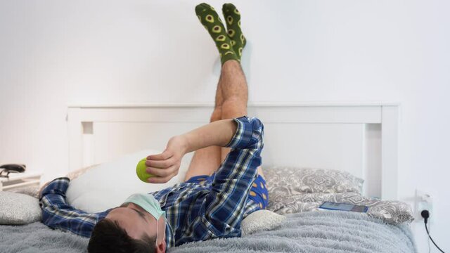 Idle young Caucasian brunette man wear medical face mask, blue shirt, panties, lies on bed, put feet on white wall. Lazy sick infected guy throws up, catches yellow tennis ball. Quarantine at home.