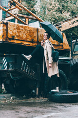 young stylish woman wearing long beige coat, white boots, black hat and backpack posing near truck crane. Trendy casual outfit. Selective focus, grain