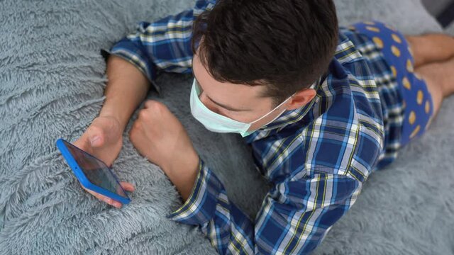 Brunette Caucasian looking man wear disposable PPE face mask, chequered flannel shirt, blue underwear lies lazily in bed, watches enjoyable video on smartphone while nodding enthusiastically. Top view